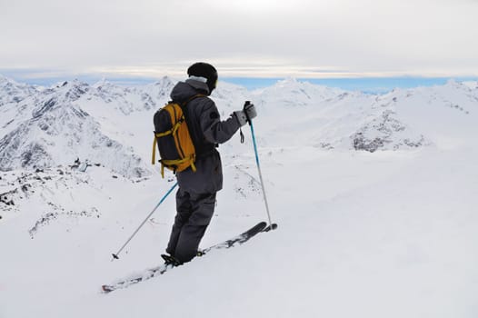 Side view of an athletic skier in a helmet and goggles with a backpack, standing on skis, holding ski poles in white snow, against the sky, enjoying a beautiful view of the mountains.