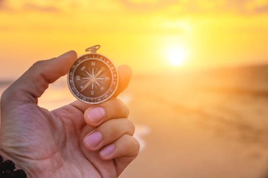 Close-up of a man's hand holding a magnetic compass against the background of the setting sun and sea