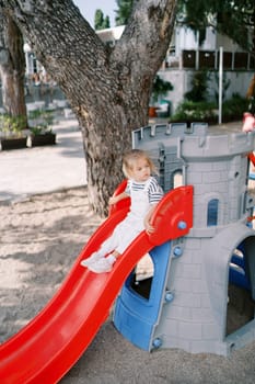 Little girl sits on top of a slide, holding the handrails and looking away. High quality photo