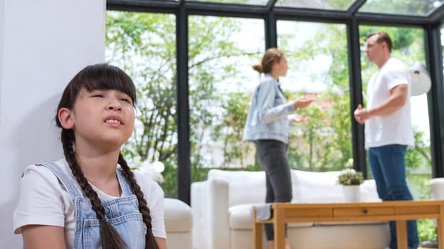 Stressed and unhappy young girl huddle in corner, cover her ears blocking sound of her parent arguing in background. Domestic violence at home and traumatic childhood develop to depression. Synchronos