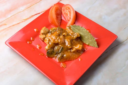 Pork stew with a bay leaf and tomato garnish on a red plate, marble background, typical food, typical mediterranean mallorcan cuisine typical from balearic islands mallorca, spain,