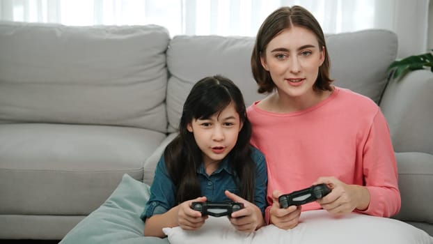 Caucasian mom and daughter sitting and playing game at living room. Happy mother spend time together with smart asian girl while holding joystick ans enjoy playing games. Lifestyle concept. Pedagogy.