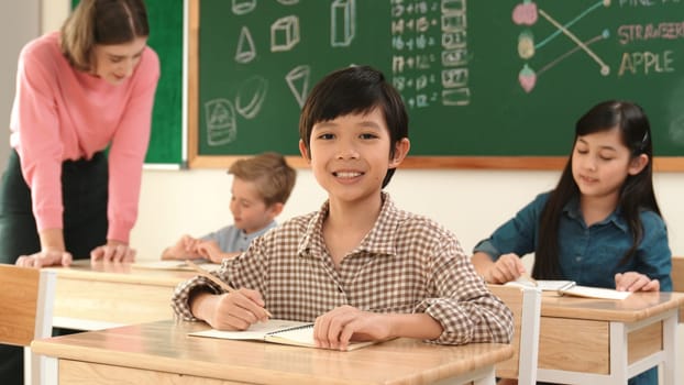 Asian smart boy smiling to camera while student writing answer in answer sheet. Multicultural student doing classwork or test while caucasian teacher checking student homework at classroom. Pedagogy.