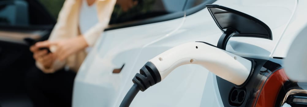 Young woman use smartphone to pay for electricity at public EV car charging station green city park. Modern environmental and sustainable urban lifestyle with EV vehicle. Panorama Expedient