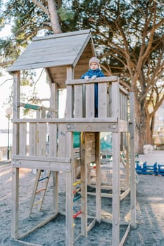 Little girl stands on the platform of a toy wooden house on the playground, leaning on the handrail and looking into the distance. High quality photo