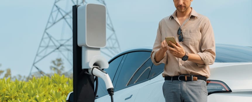 Man pay for electricity with smartphone while recharge EV car battery at charging station connected to electrical [ower grid tower for eco friendly car utilization. Panorama Expedient