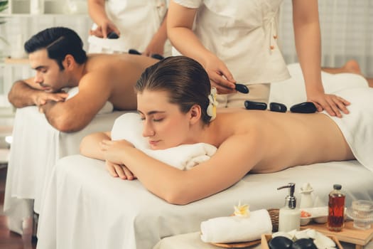 Hot stone massage at spa salon in luxury resort with day light serenity ambient, blissful couple customer enjoying spa basalt stone massage glide over body with soothing warmth. Quiescent