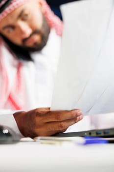 Selective focus on papers being held by a young man dressed in Arabic clothing and having a phone call. Close-up of a Muslim guy focused on examining his paperwork while speaking on a mobile device.