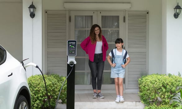 Happy little young girl learn about eco-friendly and energy sustainability as she help her mother recharge electric vehicle from home EV charging station. EV car and modern family. Panorama Synchronos