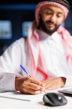 Arab man seated at his office table engrossed in his job. He is working on a minicomputer, taking detailed notes and conducting online research, showcasing a dedicated and focused approach.