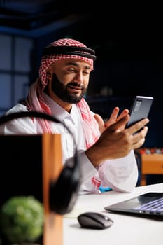 Middle Eastern entrepreneur having a serious conversation with a coworker on his mobile device in his home office. He wears Islamic clothing and has a professional demeanor.
