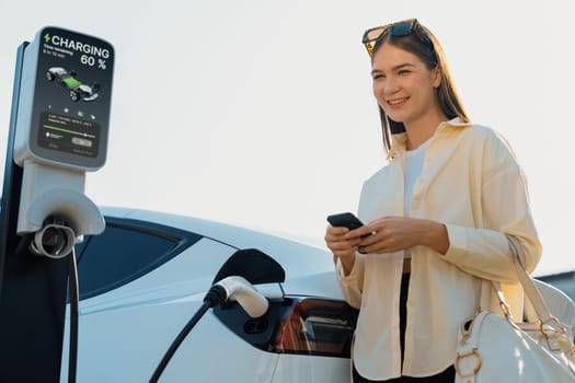 Young woman holding shopping bag and use smartphone to pay for electricity for recharging EV car battery from charging station at city mall parking lot. Modern woman go shopping by eco car. Expedient