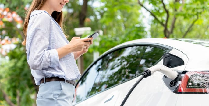 Young woman using smartphone online banking application to pay for electric car battery charging from EV charging station during vacation holiday road trip at national park or summer forest. Exalt