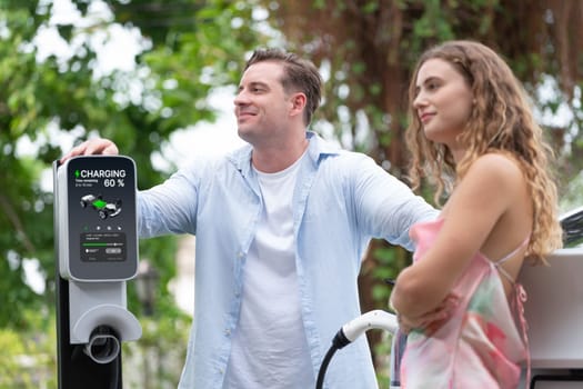 Happy and lovely couple with eco-friendly conscious recharging electric vehicle from EV charging station. EV car technology utilized as alternative transportation for future sustainability. Synchronos