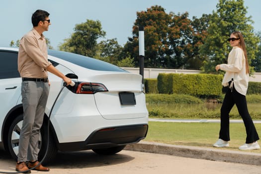 Young couple recharge electric car's battery from charging station in outdoor green city park in springtime. Rechargeable EV car for sustainable environmental friendly urban travel lifestyle.Expedient