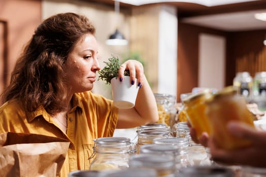 Storekeeper showing aromatic herbs to woman shopping in zero waste grocery store, inviting her to smell them. Merchant in sustainable local neighborhood shop aiding customer