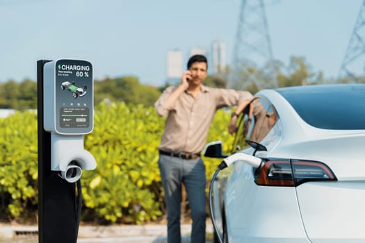 Man talking on the phone while recharge EV car battery at charging station connected to power grid tower electrical as electrical industry for eco friendly car utilization.Expedient