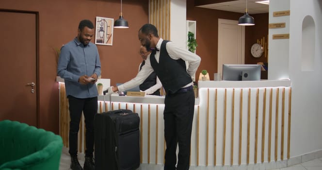 Traveller receiving room access card at front desk lobby, porter offering concierge services and helping with bags. Young hotel guest enjoying five star resort services with professional staff.