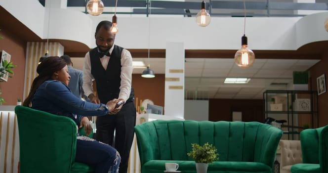 Woman paying for cup of coffee in hotel lobby, using credit card to buy drink from bar service. African american traveller making pos payment while waiting to register into room on holiday.