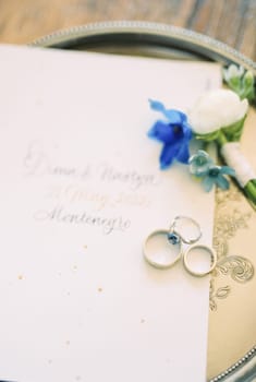 Wedding rings lie on an invitation next to a boutonniere on a tray. High quality photo