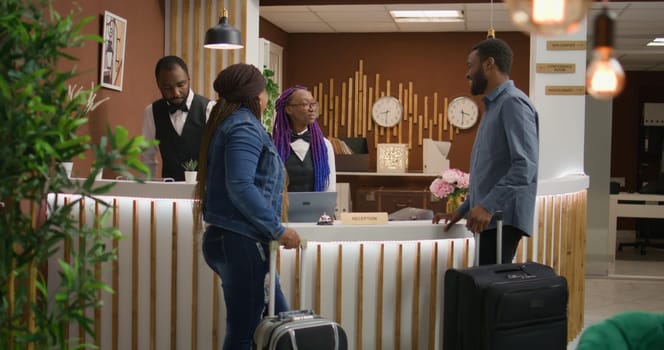 Receptionist greeting tourists at hotel, asking for personal information to verify online room booking. Couple on vacation travelling with trolley bags, arriving at front desk reception.