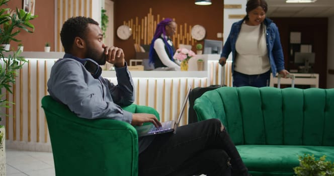 Guest looks at webpage in lobby, waiting in lounge area at hotel using laptop before registering and seeing room. African american man arriving at holiday retreat, using portable pc.