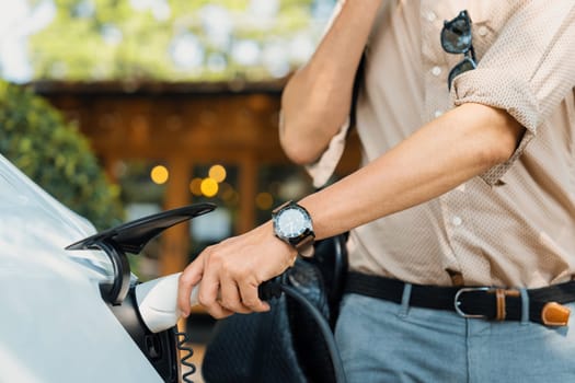 Eco friendly man and sustainable urban commute with EV electric car recharging at outdoor coffee cafe in springtime garden, green city sustainability and environmental friendly EV vehicle. Expedient