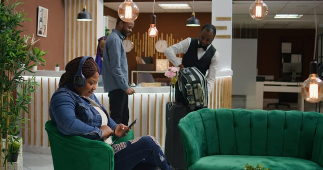 Woman listens to music on tablet in lounge area, waiting to register at front desk and start vacation. African american hotel guest using tablet and audio headset to pass time, tourism industry.