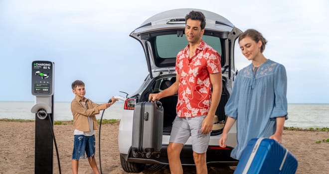 Family vacation trip traveling by the beach with electric car, lovely family taking luggage out while charging EV car battery with clean energy. Alternative family travel by eco-friendly car.Perpetual