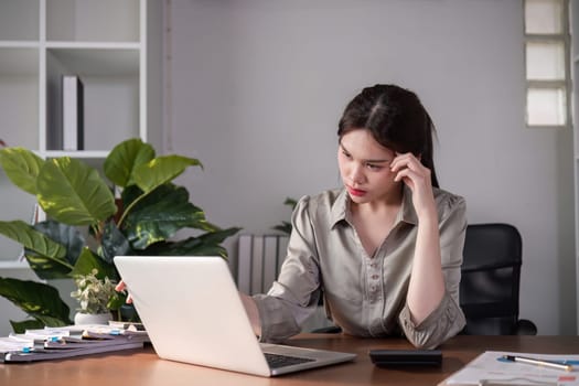 Unhappy Asian businesswoman shows stress while working in a home office decorated with soothing green plants..