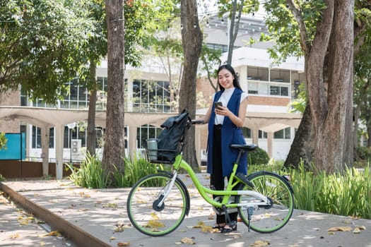 A young Asian woman commutes to work by cycling in a green city, carrying a backpack and using a reusable drinking cup to avoid harming the environment..