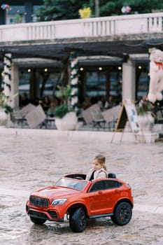 Little smiling girl drives a red toy car down the square. High quality photo