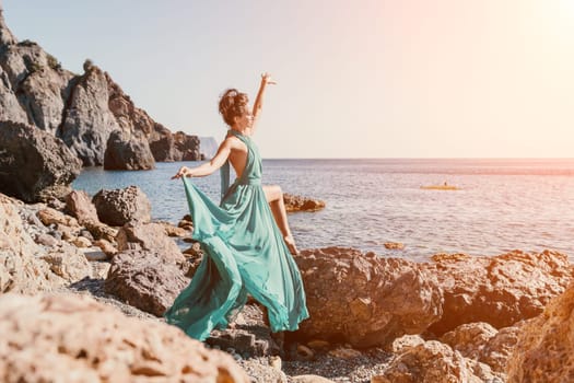 Side view a Young beautiful sensual woman in a red long dress posing on a rock high above the sea during sunrise. Girl on the nature on blue sky background. Fashion photo.