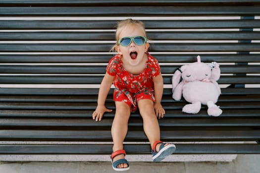 Little laughing girl sitting on a wooden bench with a pink soft toy rabbit. High quality photo