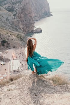 Side view a Young beautiful sensual woman in a mint long dress posing on a volcanic rock high above the sea during sunset. Girl on the nature on overcast sky background. Fashion photo