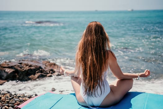 Young woman in swimsuit with long hair practicing stretching outdoors on yoga mat by the sea on a sunny day. Women's yoga fitness pilates routine. Healthy lifestyle, harmony and meditation concept.