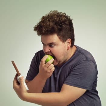 Guy, apple and chocolate in studio with choice of junk food, sweet or diet for wellness. Plus size, male person eating and decision, fruit or candy for nutrition, lose weight and healthy lifestyle.