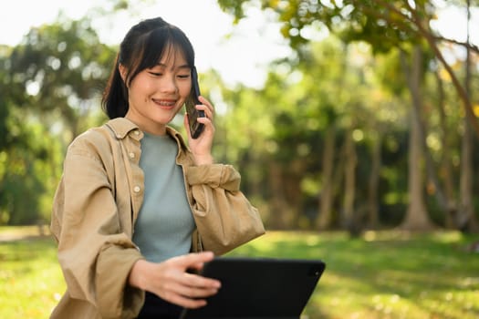 Smiling young woman talking on mobile phone and using digital tablet in the park on sunny day.