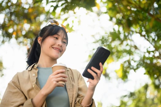 Satisfied Asian woman drinking coffee from paper cup using mobile phone in the park.