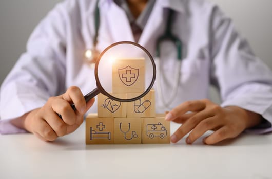 Doctor holding magnifying glass against stacked wooden cubes with medical insurance icon.