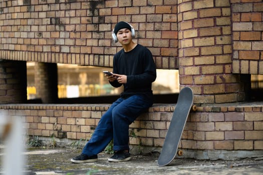 Full length of Asian man in stylish clothes sitting with skateboard at city street and using mobile phone.