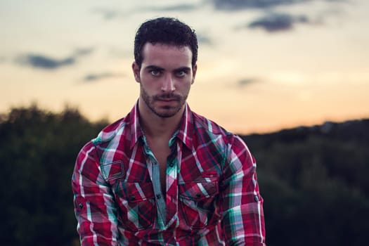 A man wearing a red and green plaid shirt is standing outdoors. He is looking straight ahead, with a relaxed expression on his face, against a simple background.