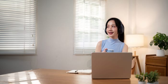 Happy young woman asian in headphones and laptop making notes, girl student talking by video conference call, online training.