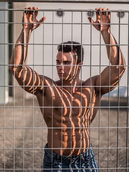 A shirtless muscular man standing behind a metal fence, with his hands in the air. The man appears to be in distress or surrendering. The background is blurred, focusing on the mans actions.