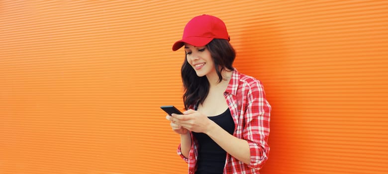 Portrait of happy smiling young woman with mobile phone in summer red baseball cap on colorful orange background