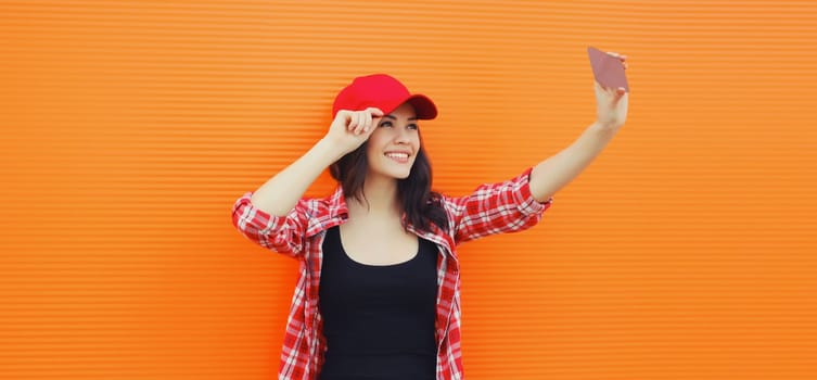 Summer portrait of happy cheerful smiling young woman taking selfie with mobile phone in red baseball cap on colorful orange background on city street