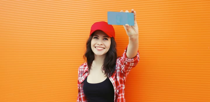 Summer portrait of happy cheerful smiling young woman taking selfie with mobile phone in red baseball cap on colorful orange background on city street