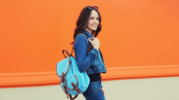 Summer portrait of happy smiling brunette young woman with backpack walking along city street, orange background