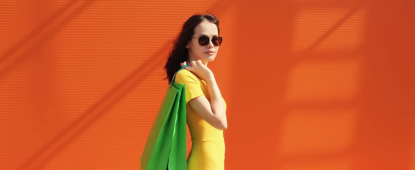 Portrait of stylish beautiful young woman posing with shopping bags on orange background in city