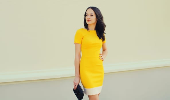 Beautiful young brunette woman holding handbag clutch posing in yellow dress on city street